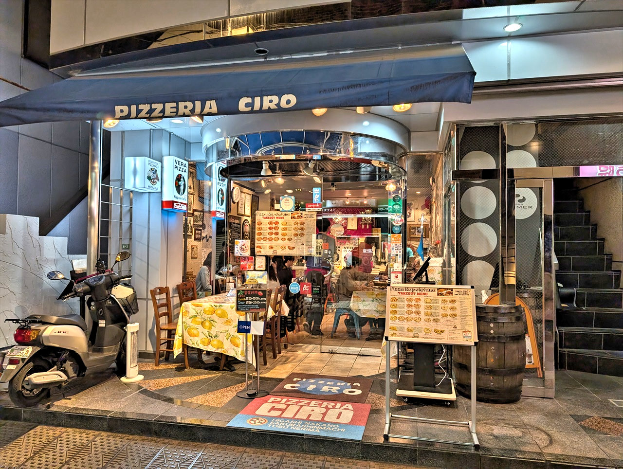 夜の外観。「PIZZERIA CIRO」の青いひさしと円形の入口アーチ、店内に並ぶ木製テーブルと椅子、壁面に貼られた複数のメニュー看板が見える